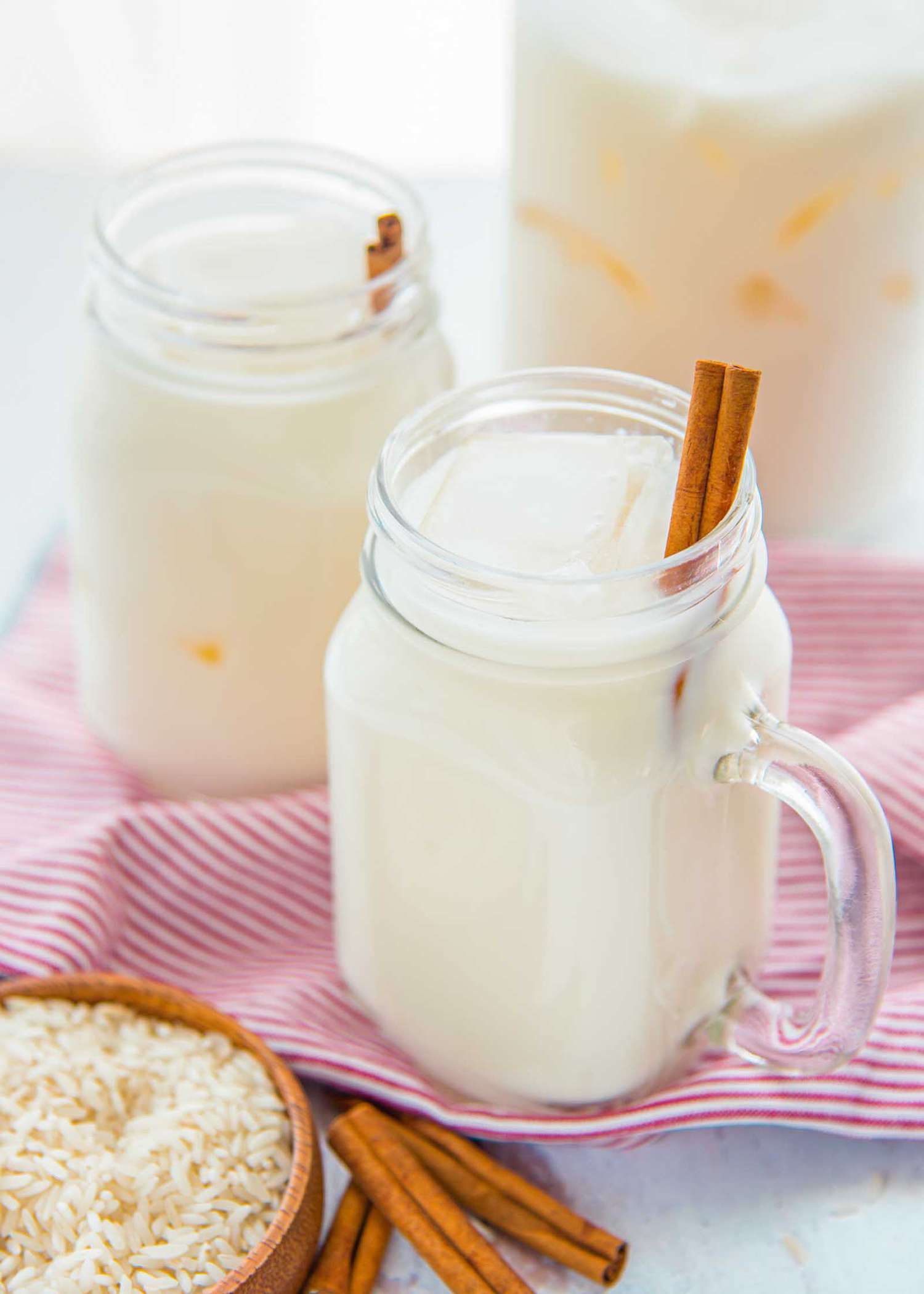 Easy homemade horchata over ice with a cinnamon stick are in a mason jar glass. A second identical glass is behind it and a pink striped linen is underneath. In the upper right hand corner is a pitcher of creamy horchata. A small bowl of rice and cinnamon sticks are in the lower left hand corner.