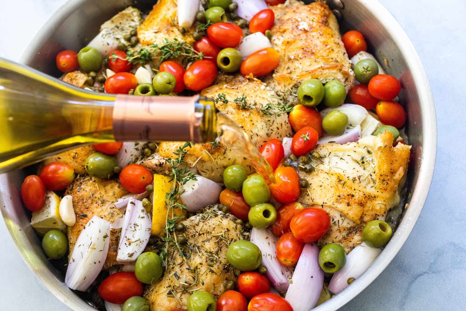 Adding wine to chicken with shallots, garlic, cherry tomatoes, green olives, lemon wedges, capers, and thyme sprigs in pan