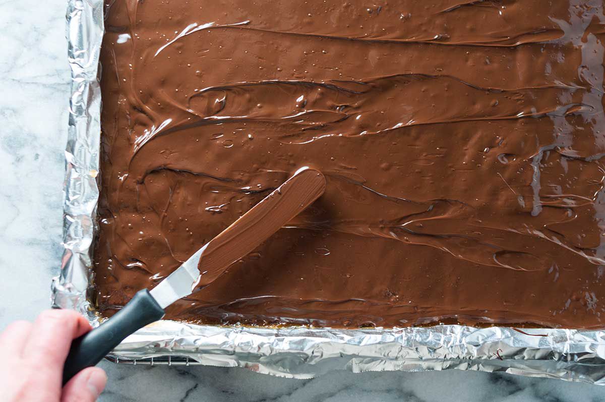 Overhead of a butter knife completely covering the baked saltines with a melted chocolate layer on a foiled baking sheet