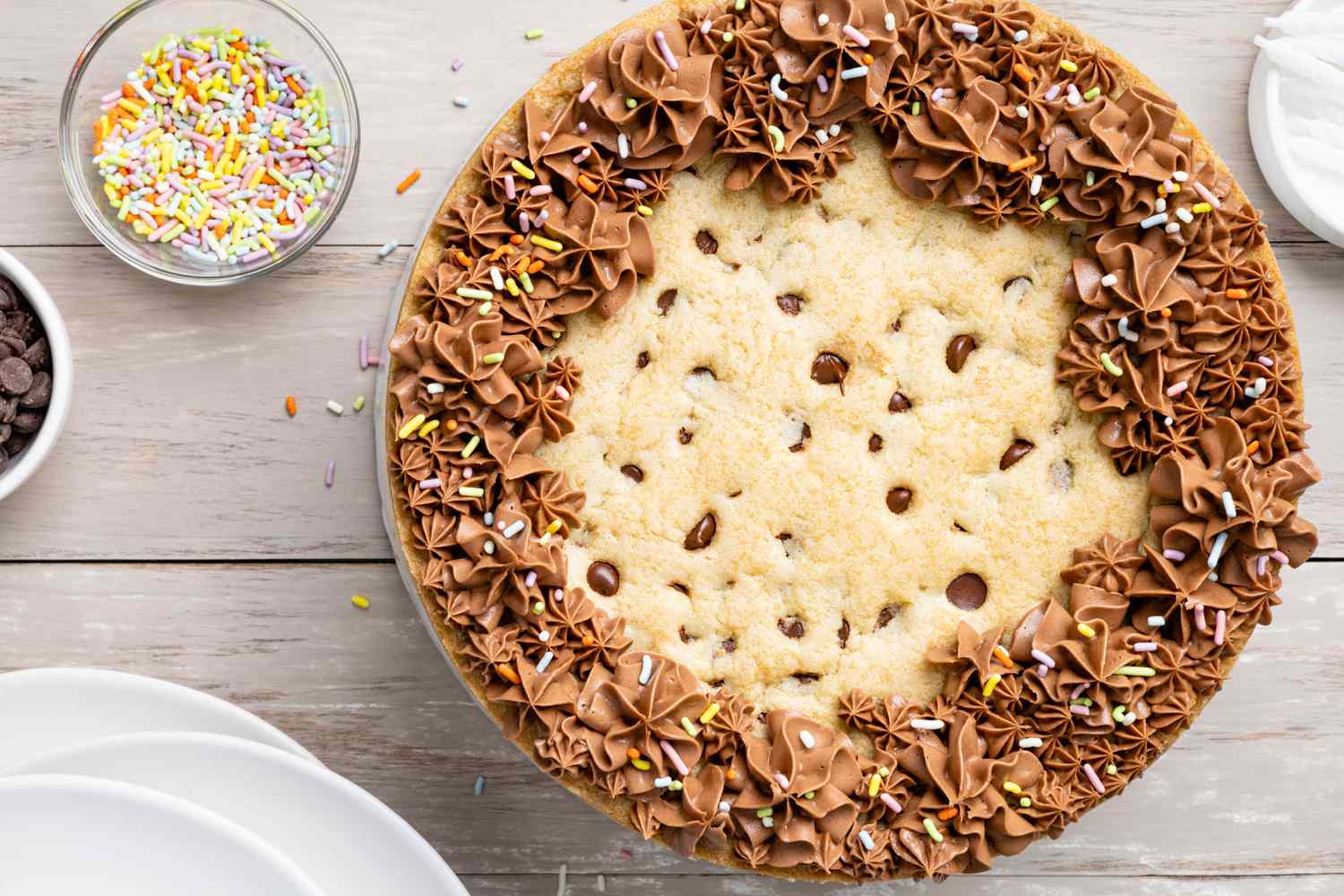 Chocolate Chip Cookie Cake Surrounded by Plates and Small Bowls with Sprinkles and Chocolate Chips