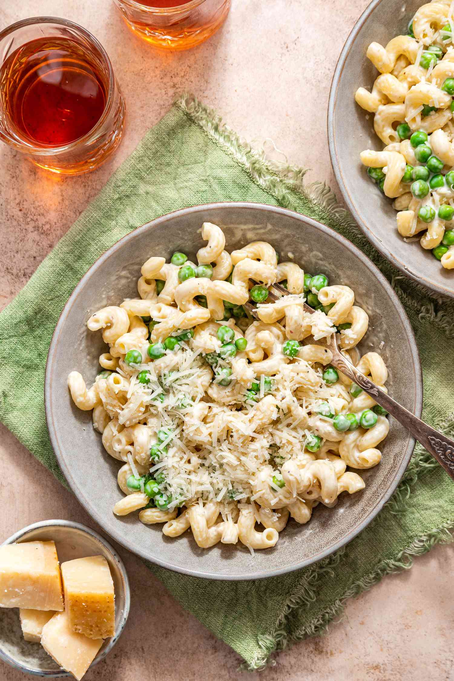 Bowl of Creamy Cavatappi Topped With Shredded Parmesan With a Fork and Sitting on an Olive Kitchen Towel. In the Surroundings, Another Bowl With a Serving, a Small Bowl With Blocks of Parmesan, and a Two Glasses.