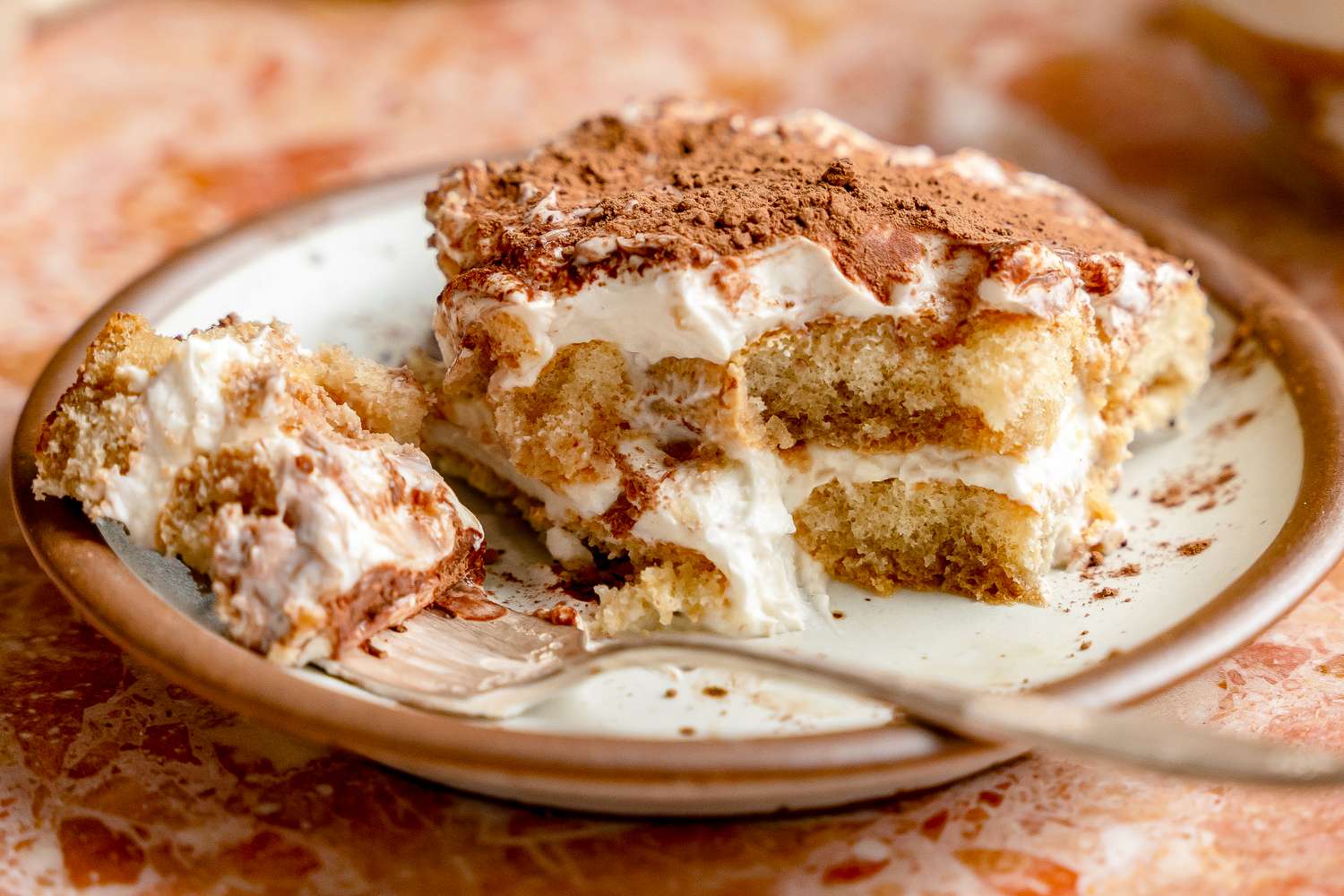 A plate with a slice of layered tiramisu topped with cocoa powder and a fork holding a portion of it