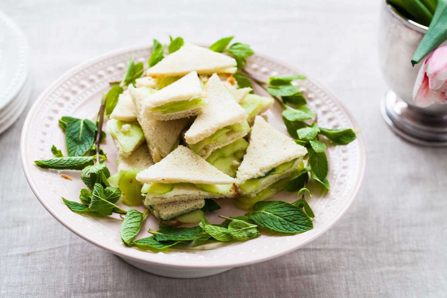 A cake plate with triangle cut cucumber dill sandwiches stacked on top. Herbs encircle the sandwiches.