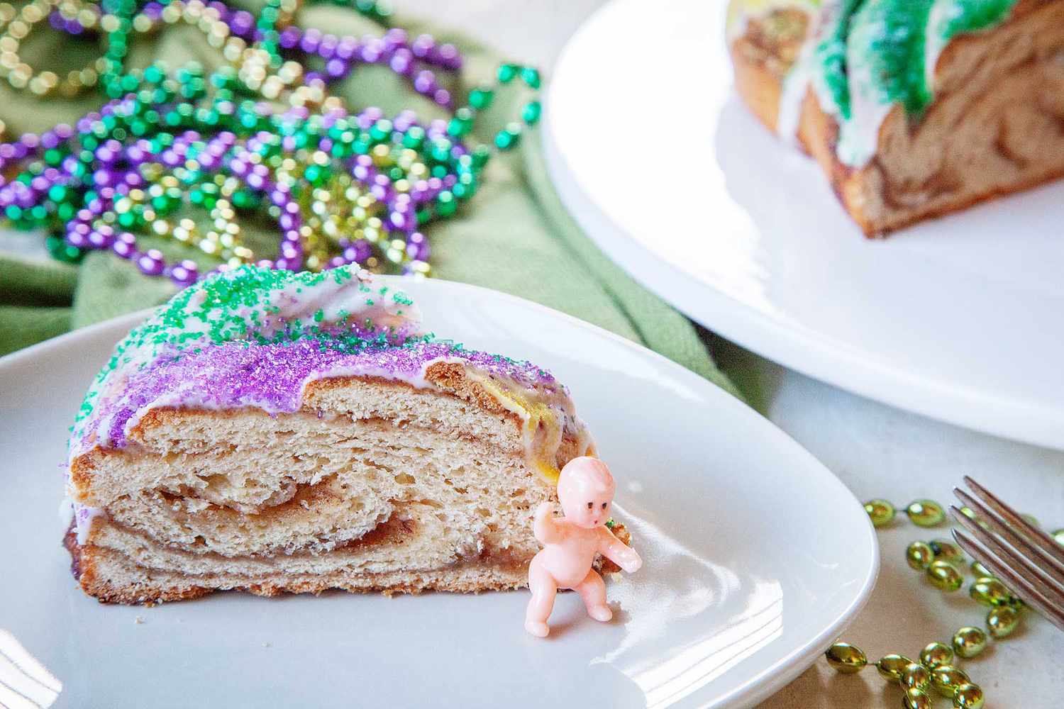Horizontal view of a slice of homemade king cake with a small baby figurine on the plate. Mardi Gras beads and a partial view of the rest of the cake are in the background.