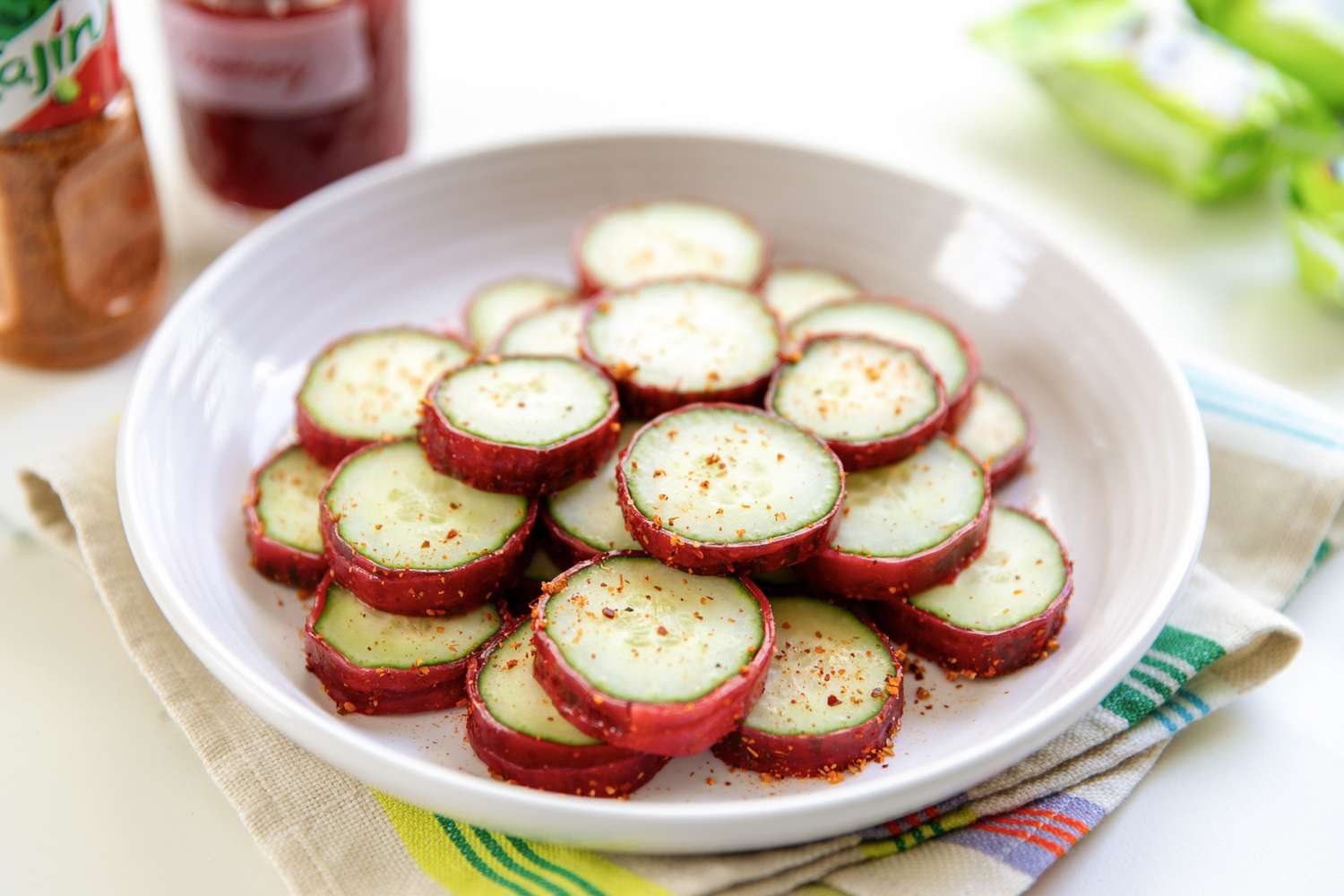 Sliced cucumbers coated in Tajin spice arranged in a white bowl on a colorful towel