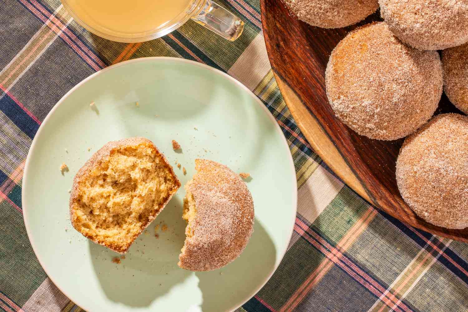 A halved apple cider donut muffin on a plate, and in the surroundings, a cup of apple cider vinegar and more muffins stacked on a plate