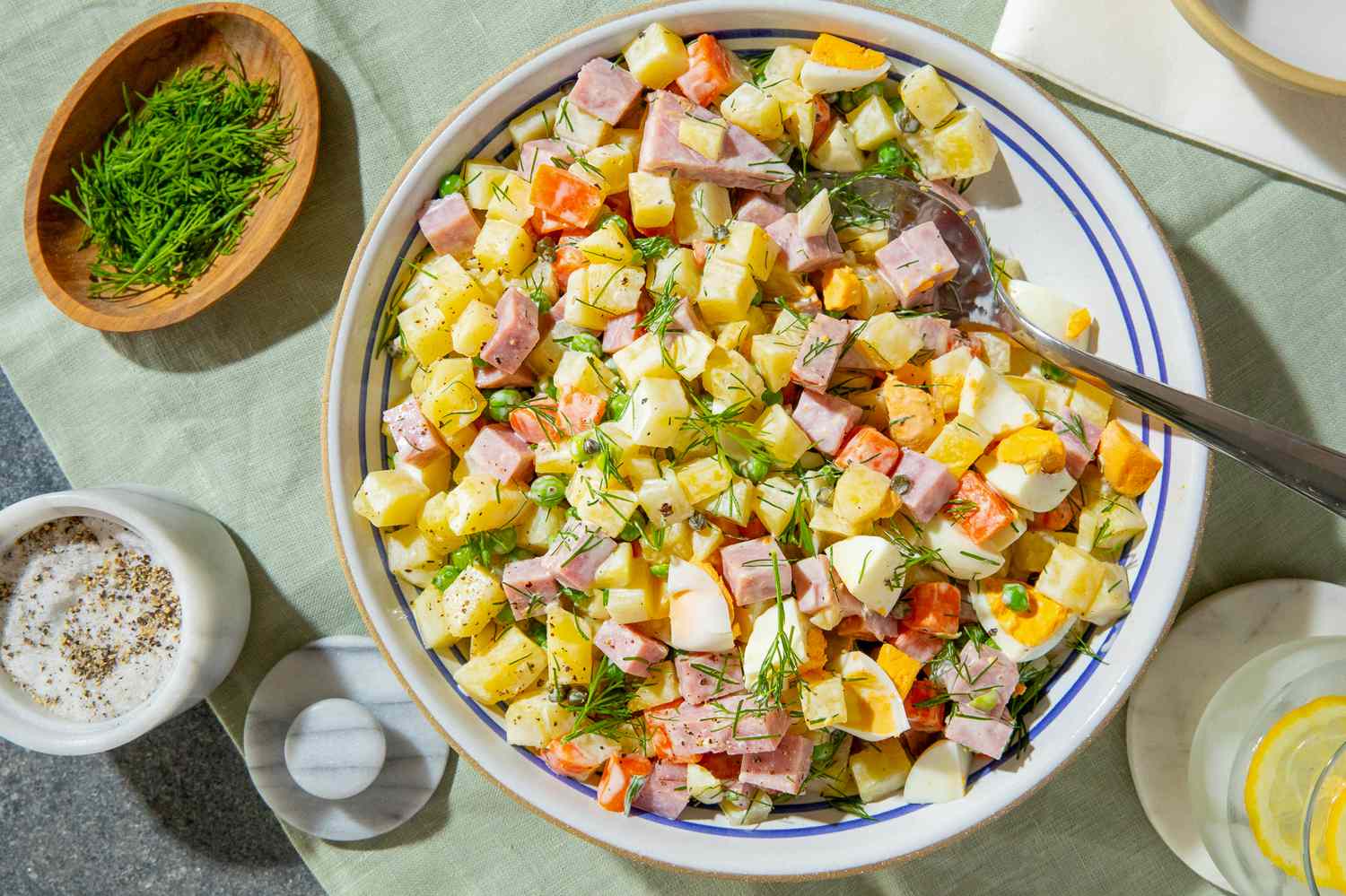bowl of olivier potato salad on a table with a small bowl of fresh dill, a countainer with salt and pepper, and a glass of lemon water 