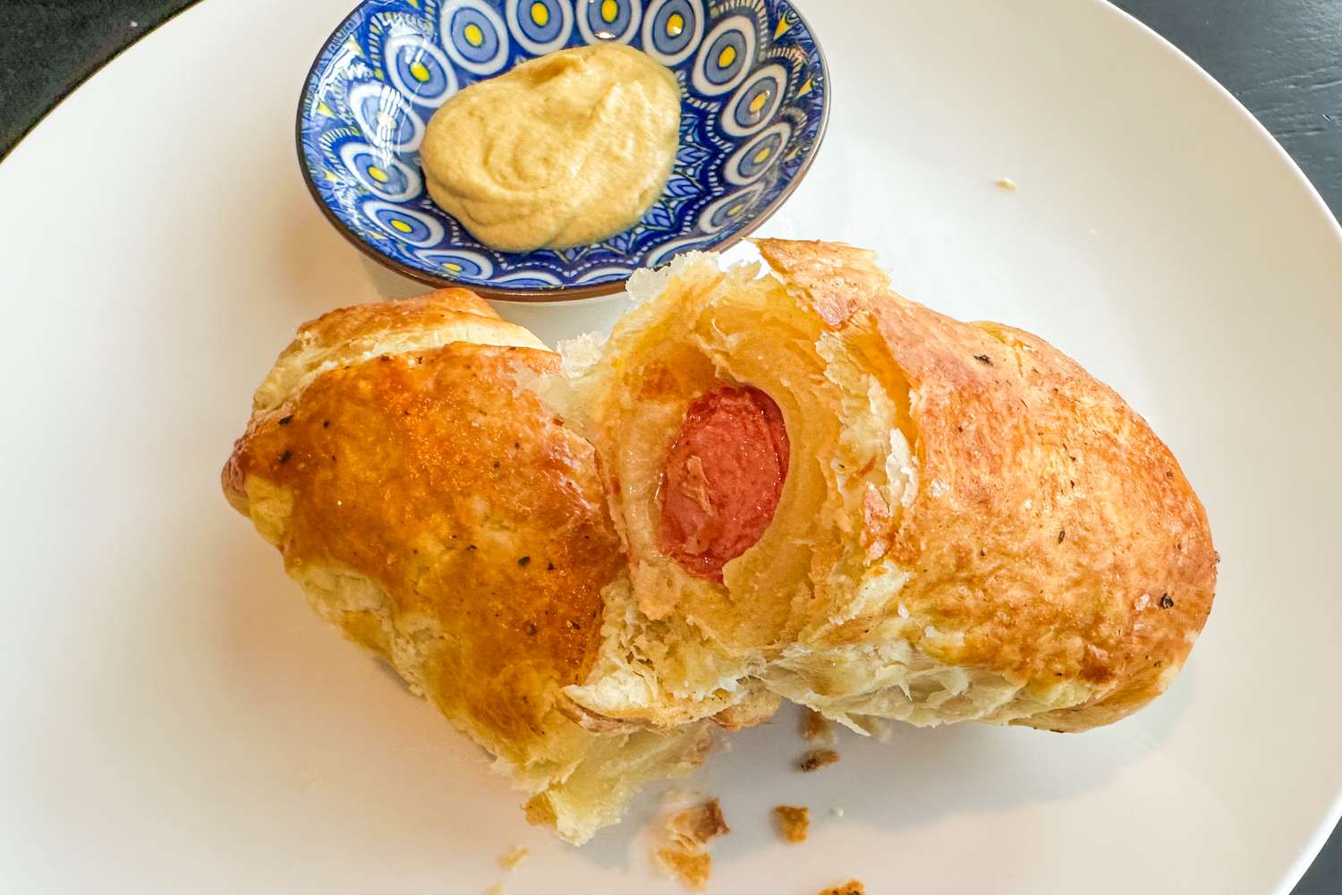 A hotdog wrapped in puff pastry split in half on a plate with a side of mustard
