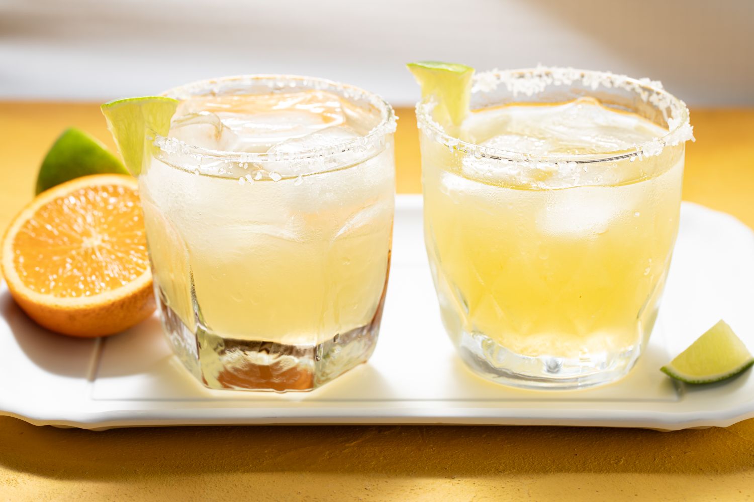 Two margarita cocktails served in glasses with slices of lime displayed on a tray with lemon and lime wedges nearby