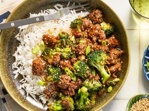 Ground beef and broccoli stir fry over rice in a bowl at a table setting with a glass of water, a bowl of sliced scallions, and a bowl of sesame seeds