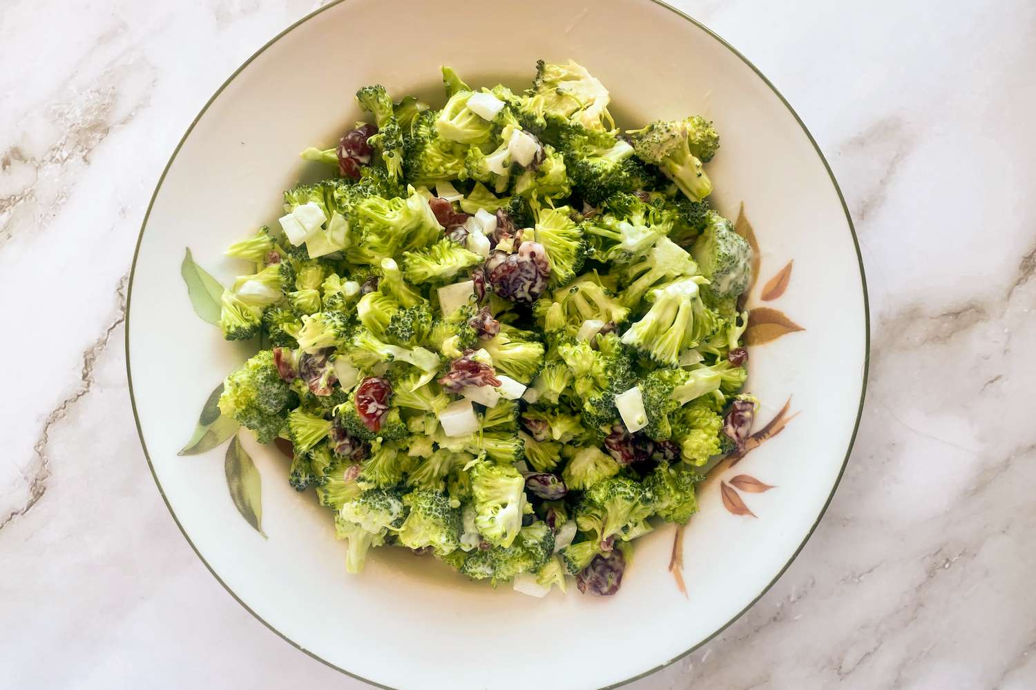 A bowl of broccoli salad with cranberries and onions on a marble surface
