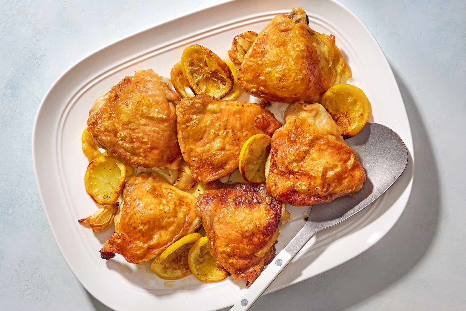 Overhead view of a white serving platter of lemon chicken thighs and lemon slices with a large serving spoon