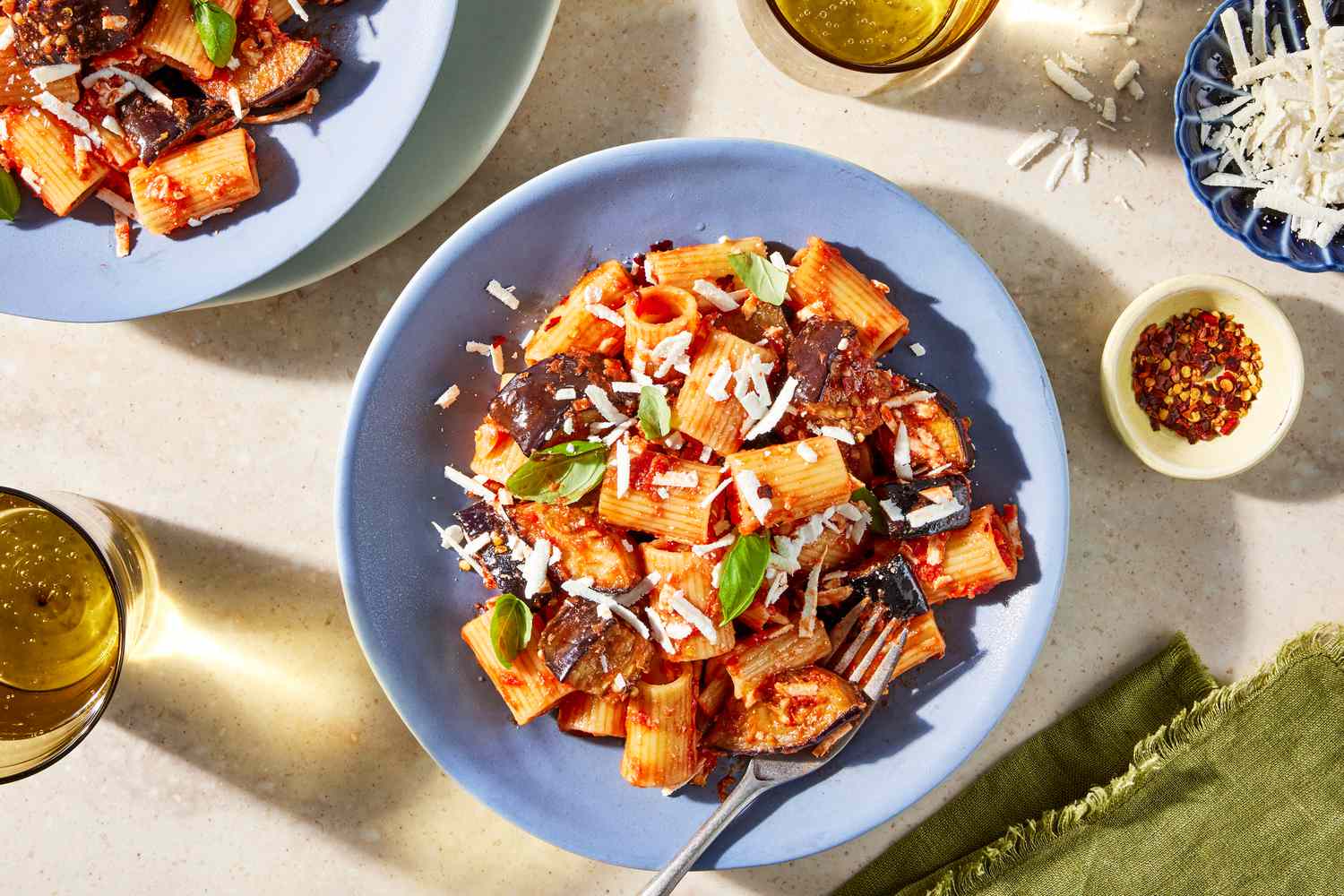 pasta alla norma on a plate at a table setting with another serving on a plate, a bowl of shredded cheese, a bowl of crushed peppers, a table napkin, and two glasses of wine