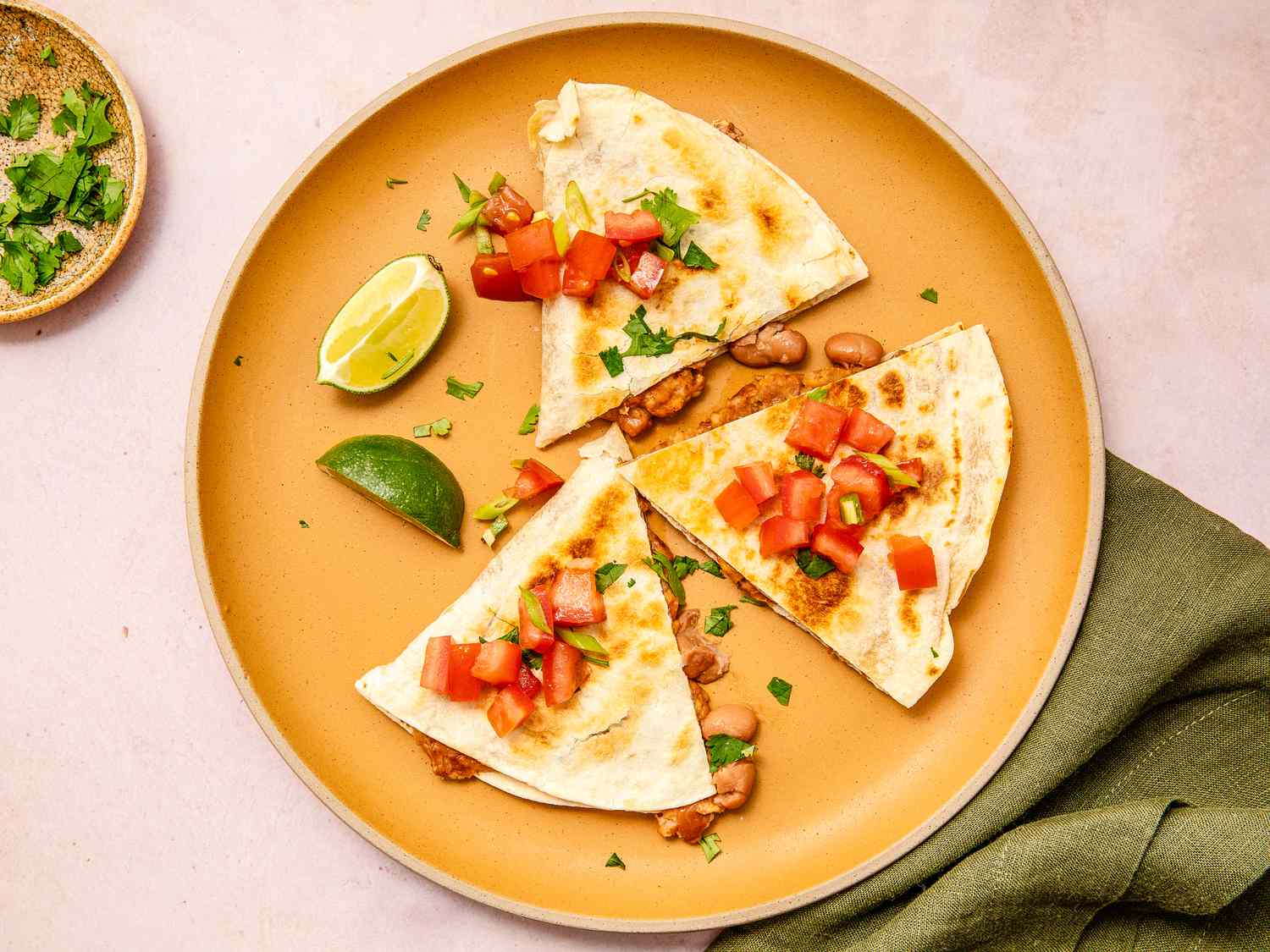 Three slices of a quesadilla on a plate along with lime slices at a table setting with a table napkin and small plate of cilantro 