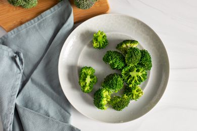 Blanched Broccoli on a Plate Surrounded by a Kitchen Towel and a Cutting Board with Heads of Broccoli for How to Blanch Broccoli
