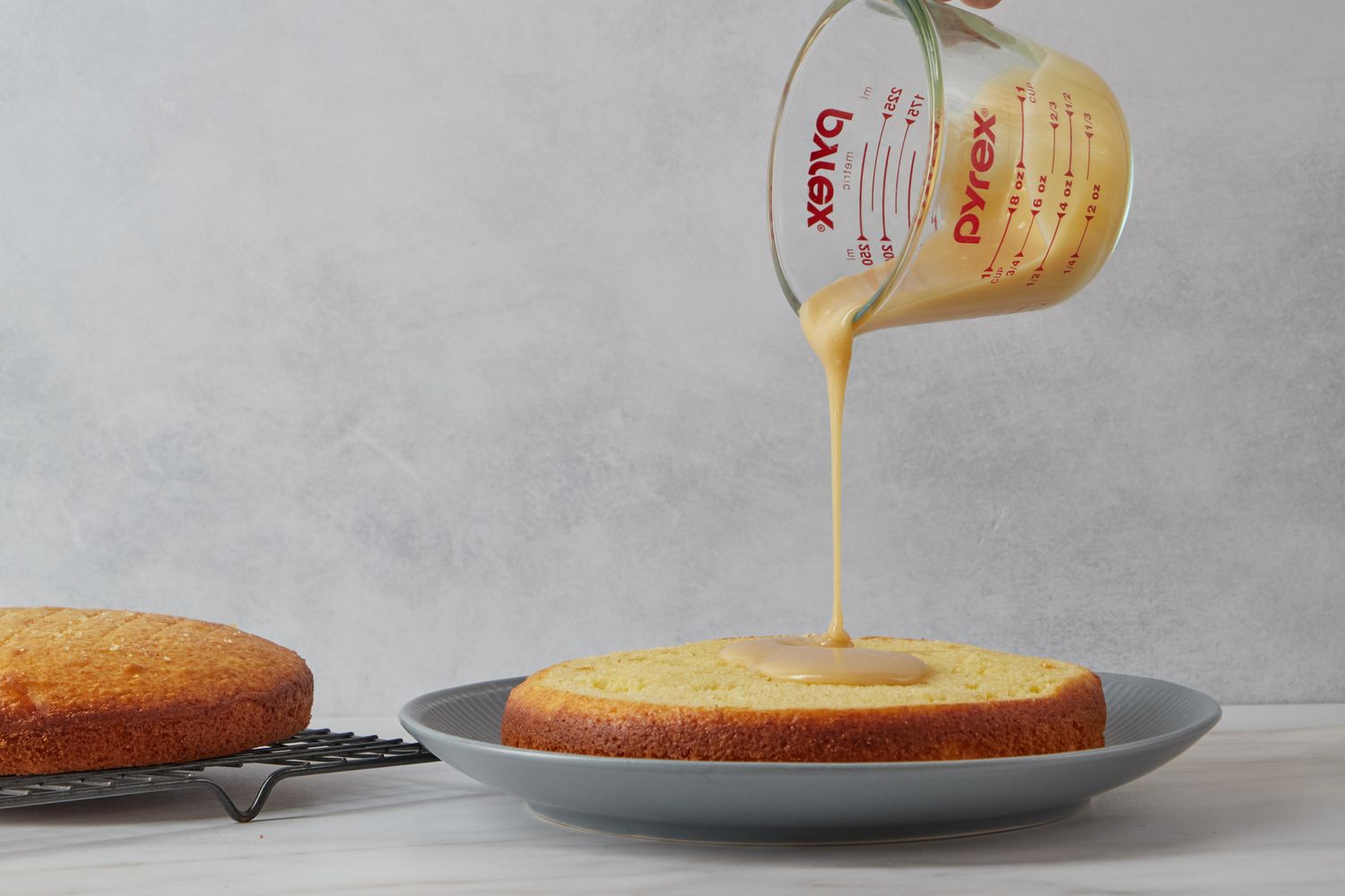 Pouring liquid onto a round cake layer from a measuring cup with another cake layer cooling on a rack in the background for Reese Witherspoon's Sour Cream Cake