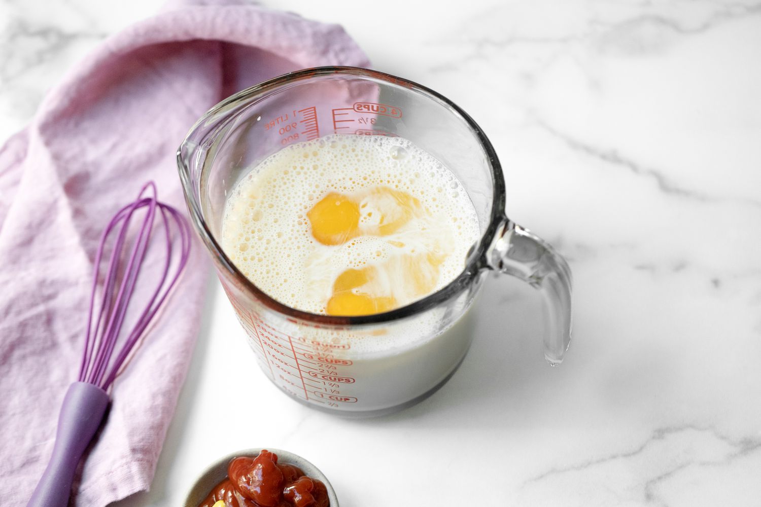 Eggs Added to Glass Measuring Cup Filled with Evaporated Milk Next to a Whisk for Caribbean Macaroni Pie Recipe