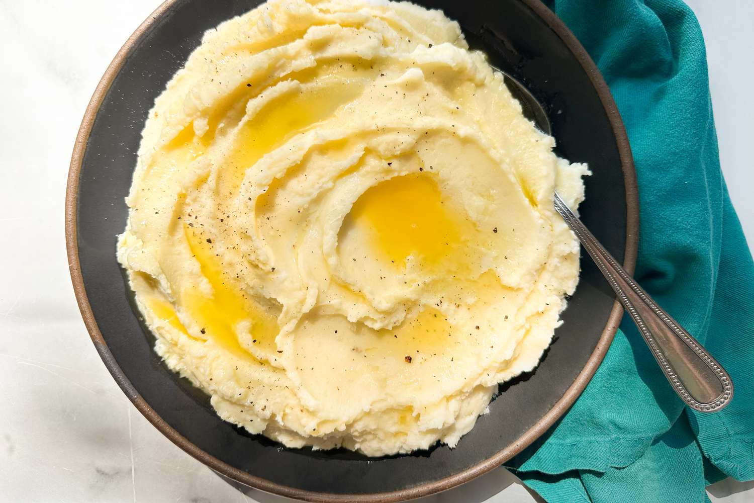Overhead view of a large, dark colored serving bowl of mashed potatoes topped with butter and with a serving spoon on the side 