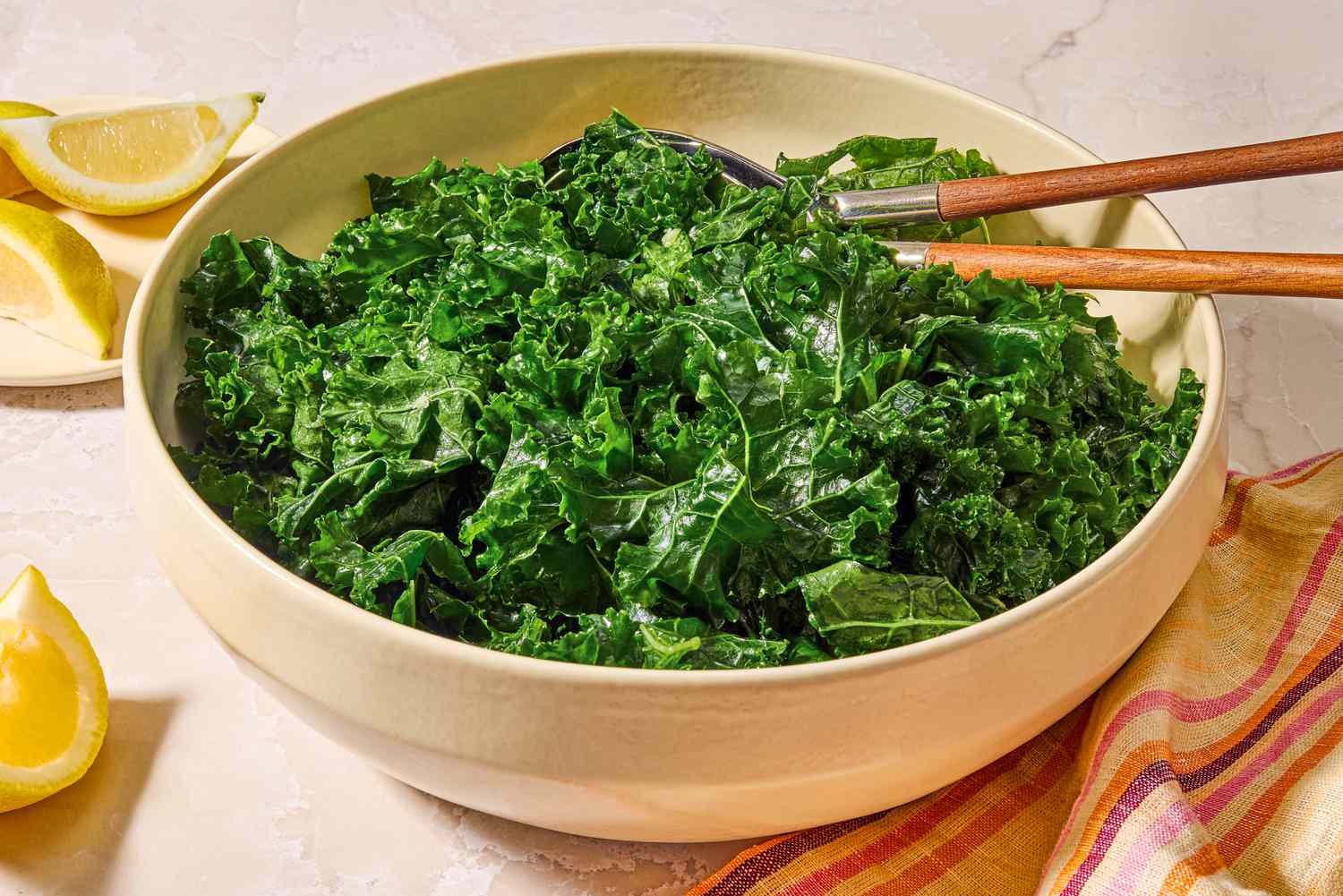 Angled view of a large bowl of kale salad with serving spoons next to lemon slices and a cloth napkin