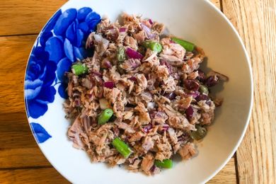 A bowl containing a mixture of tuna chopped onions green vegetables and herbs placed on a wooden table