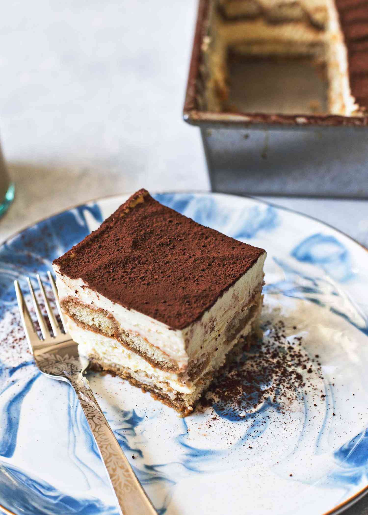 A slice of tiramisu on a plate with the pan of tiramisu in the background