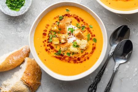 Bowls of Mercimek Corbasi (Turkish Lentil Soup) Topped with Greek Yogurt, Croutons, Pul Bibir Butter, and Parsley, and Next to It, a Small Bowl of Parsley, Slices of Bread, and Two Spoons