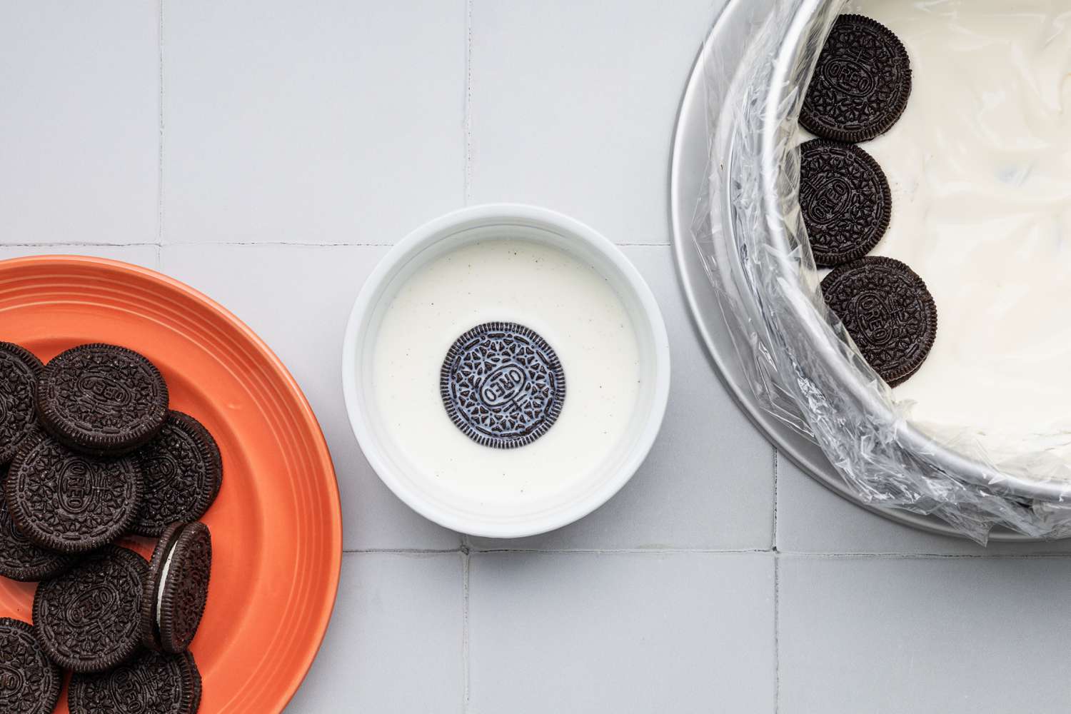 Assembling First Layer of Oreo Ice Cream Cake (L to R): Plate With Oreo Cookies, Bowl of Milk With an Oreo Cookie Soaking, Plastic Wrapped Cake Pan With Milk Dipped Cookies Added on Top of the No Churn Ice Cream