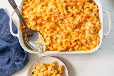 Overhead view of a white baking dish of macaroni and cheese with a serving spoon along with a blue cloth napkin and small plate of the macaroni all on a white background