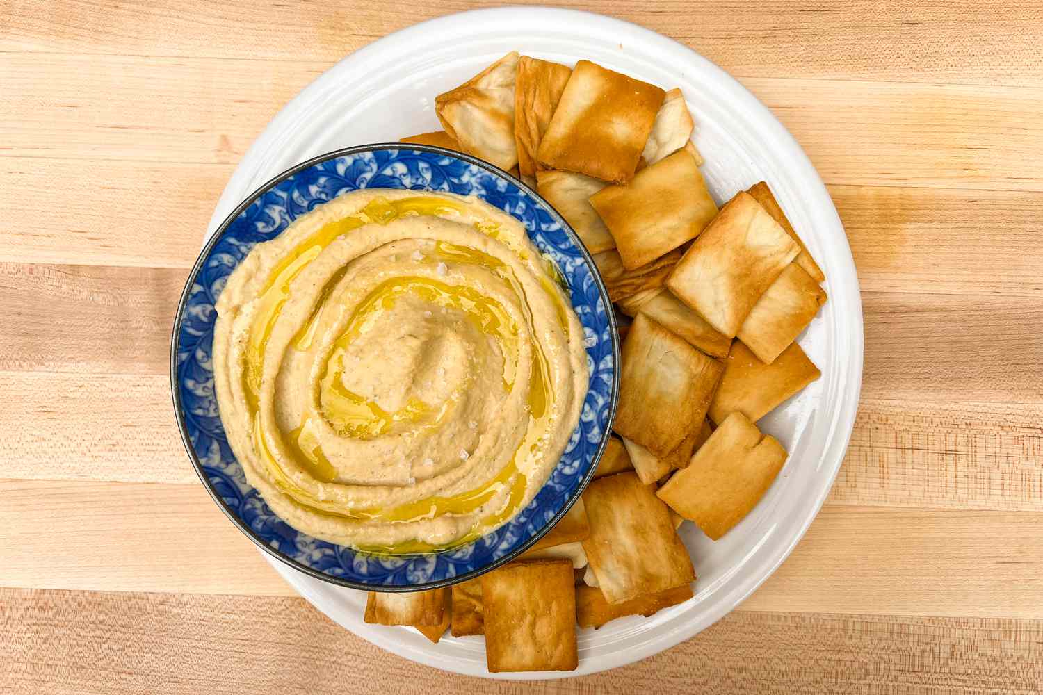 Hummus in a bowl, served with pita chips on a white plate.