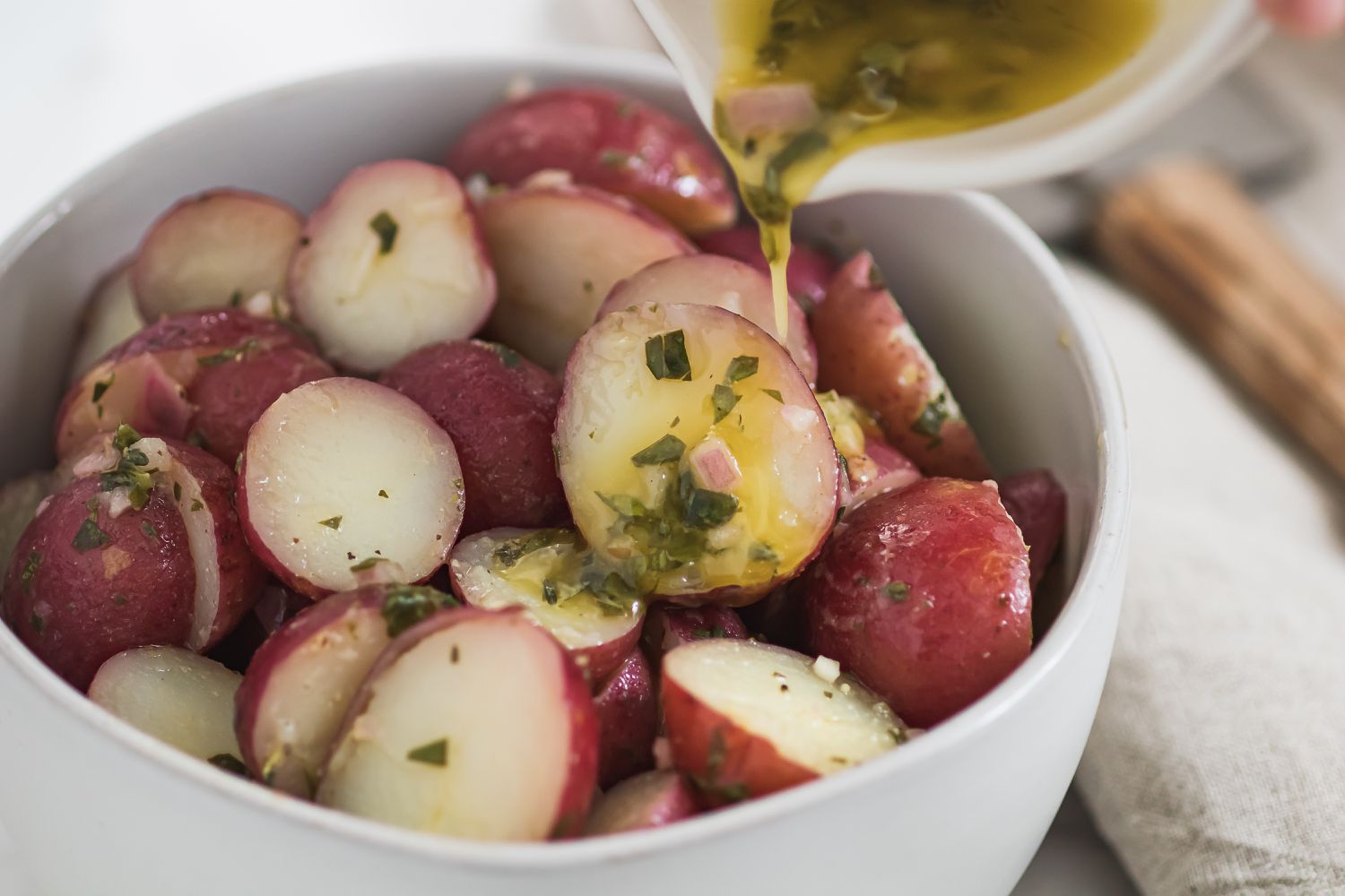 Adding viniagrette to a bowl of halved red potatoes for a nicoise salad recipe.