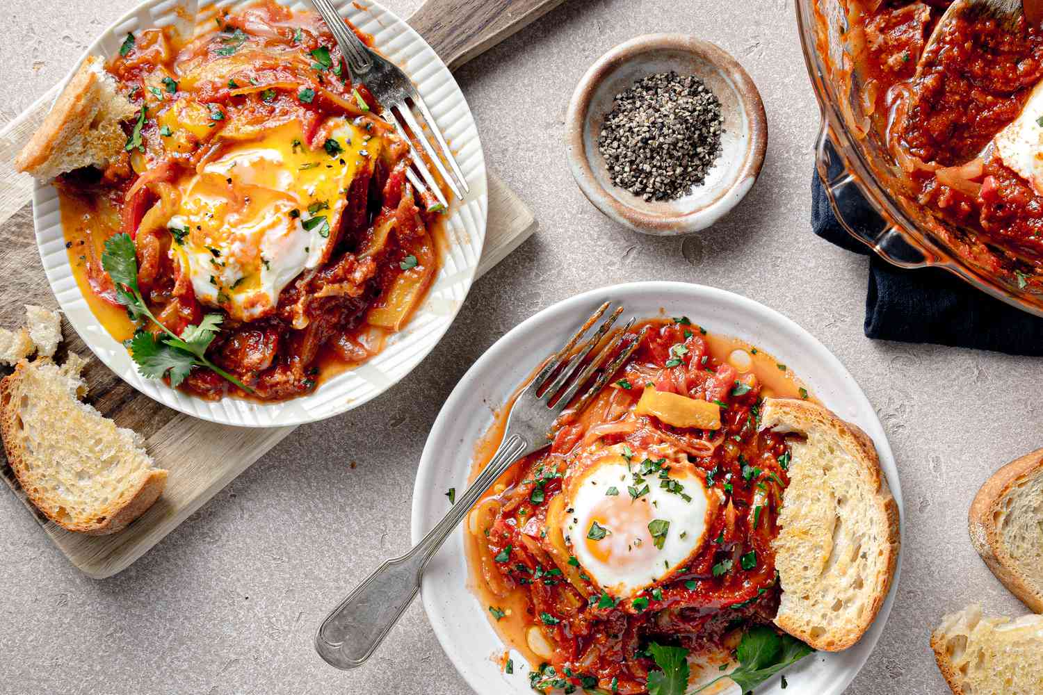 Serving of Microwave Shakshuka with a Piece of Toasted Bread on a Small Plate, and in the Surroundings, More Bread on the Counter, Another Serving of Shakshuka on Another Plate, a Small Saucer with Cracked Pepper, and More Shakshuka in a Pyrex Dish