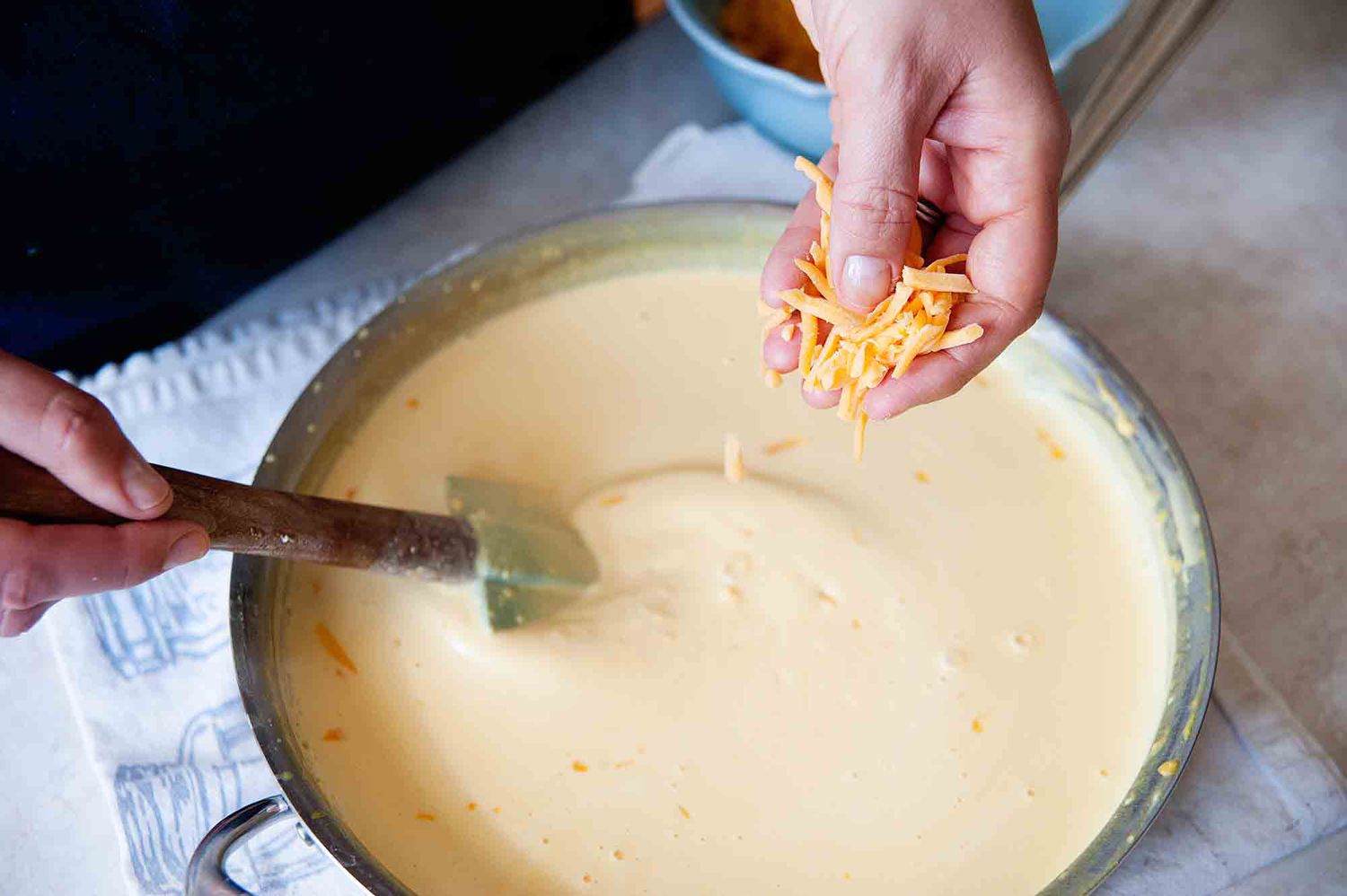 Creamy Mac and cheese recipe - hand sprinkling shredded cheddar cheese into a cheese sauce and stirring it together.