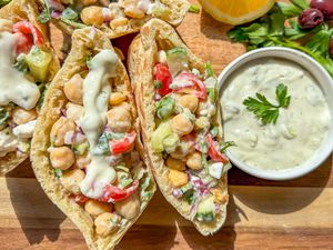 Greek salad pockets next to a some olive, fresh herbs, a halved lemon, and small bowl of tzatziki, all on a cutting board 