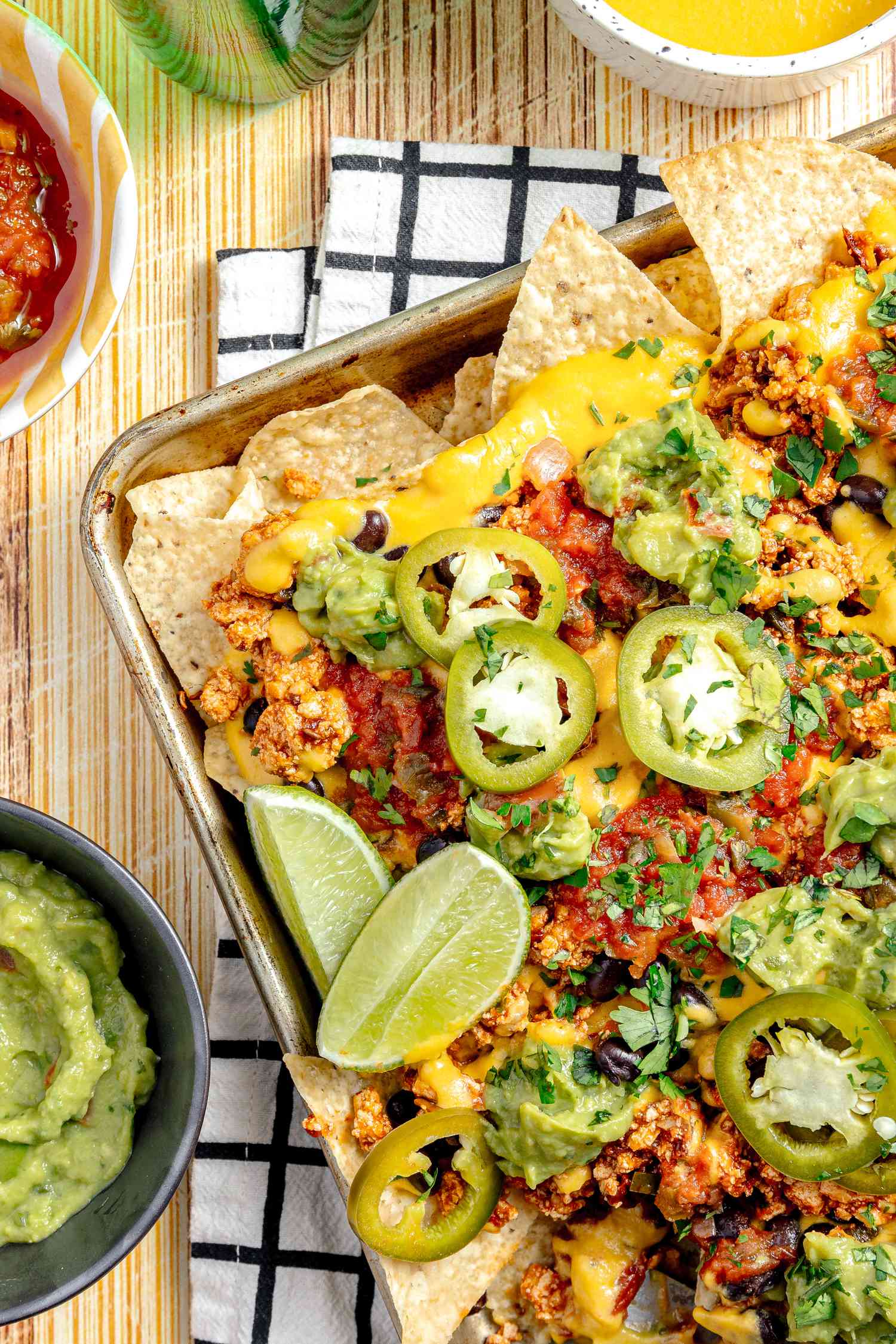 Baking Sheet of Vegan Nachos (Nachos Topped with Salsa, Queso, Pickled Jalpenos, and Two Lime Wedges) Surrounded by Bowls of Guacamole, Salsa, and Queso