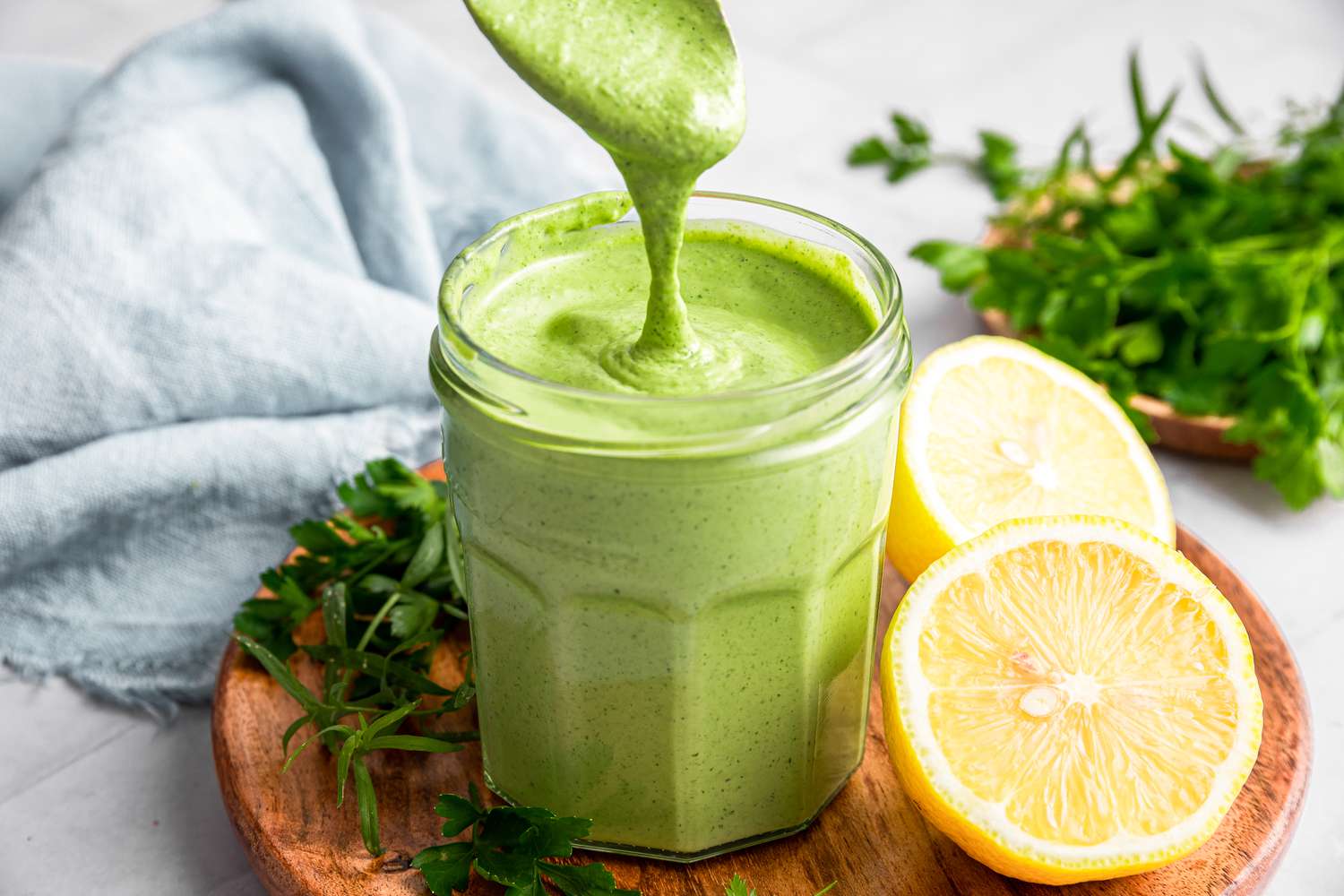 Green Goddess Dressing Dripping from a Spoon into a Jar with More Dressing. Jar is on a Wooden Plate with Herbs and Halved Lemons