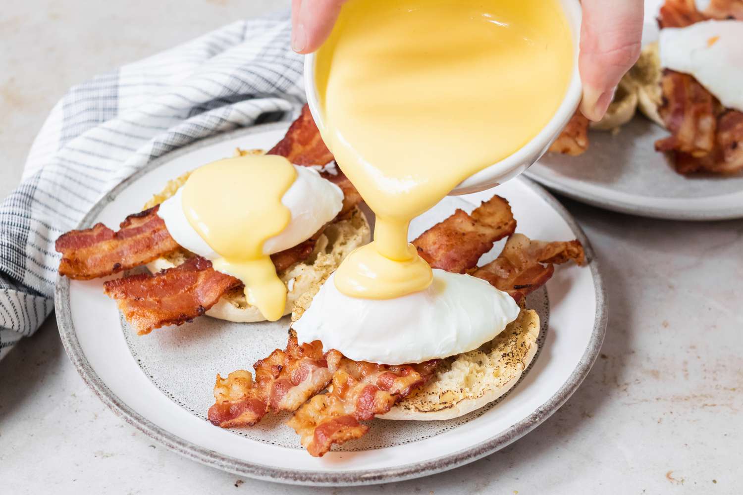 English muffins with bacon, poached eggs, and being topped with hollandaise sauce