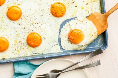 Wooden Spatula Lifting a Sheet Pan Fried Egg From a Baking Pan, and Next to the Sheet Pan, a Stack of Plate With Utensils and a Kitchen Towel