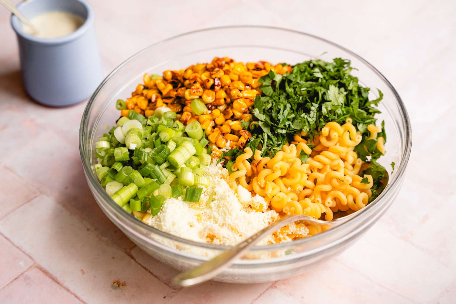 A bowl containing pasta green onions cilantro roasted corn and cheese with a dressing container in the background