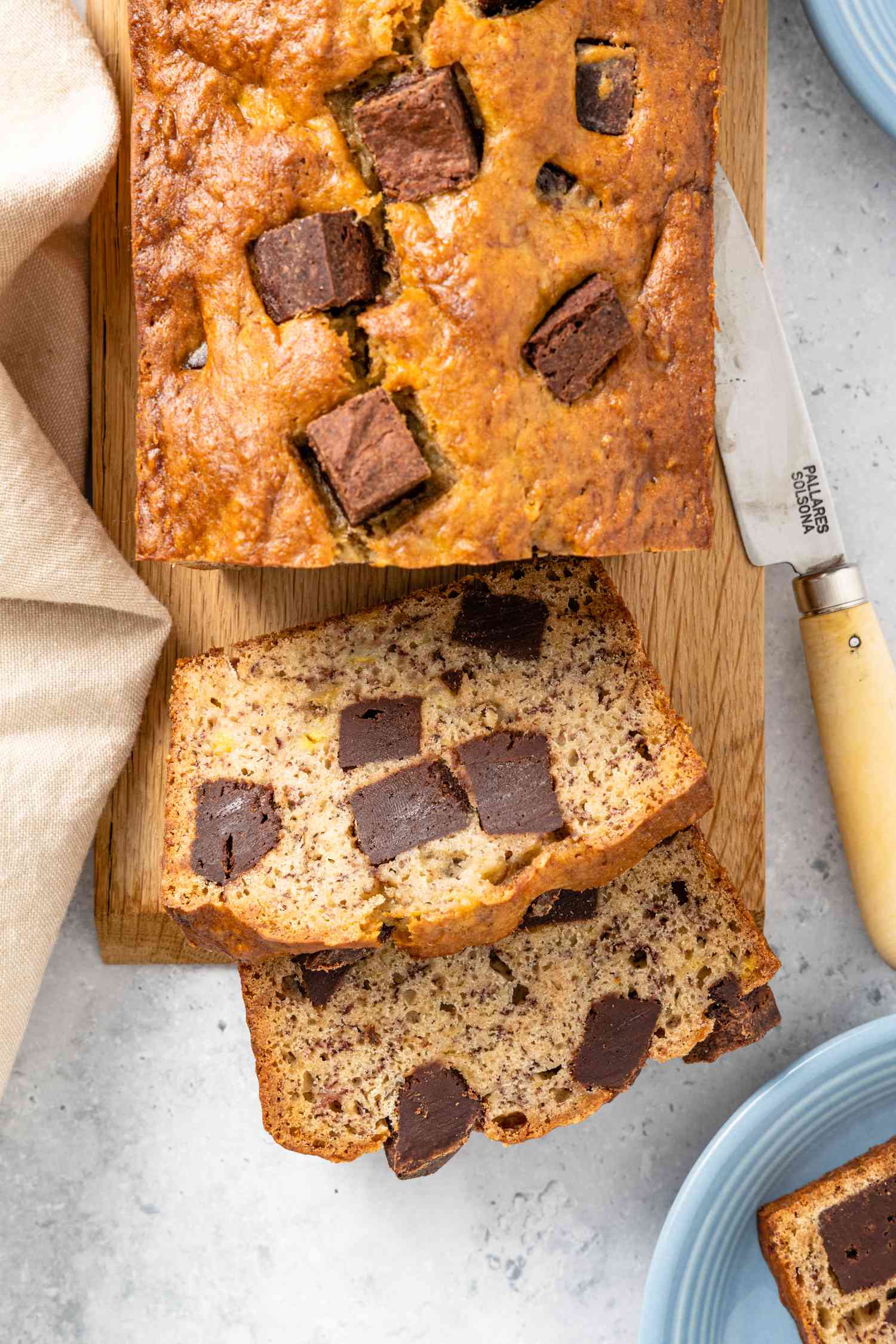 On a Wooden Board, Loaf of Brownie Banana Bread with Slices Cut Off, and in the Surroundings, a Plate with a Slice, a Knife, and a Kitchen Towel