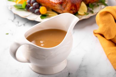 Angled view of a white gravy boat full of brown gravy next to a platter of turkey and an orange fabric napkin all on a marble counter