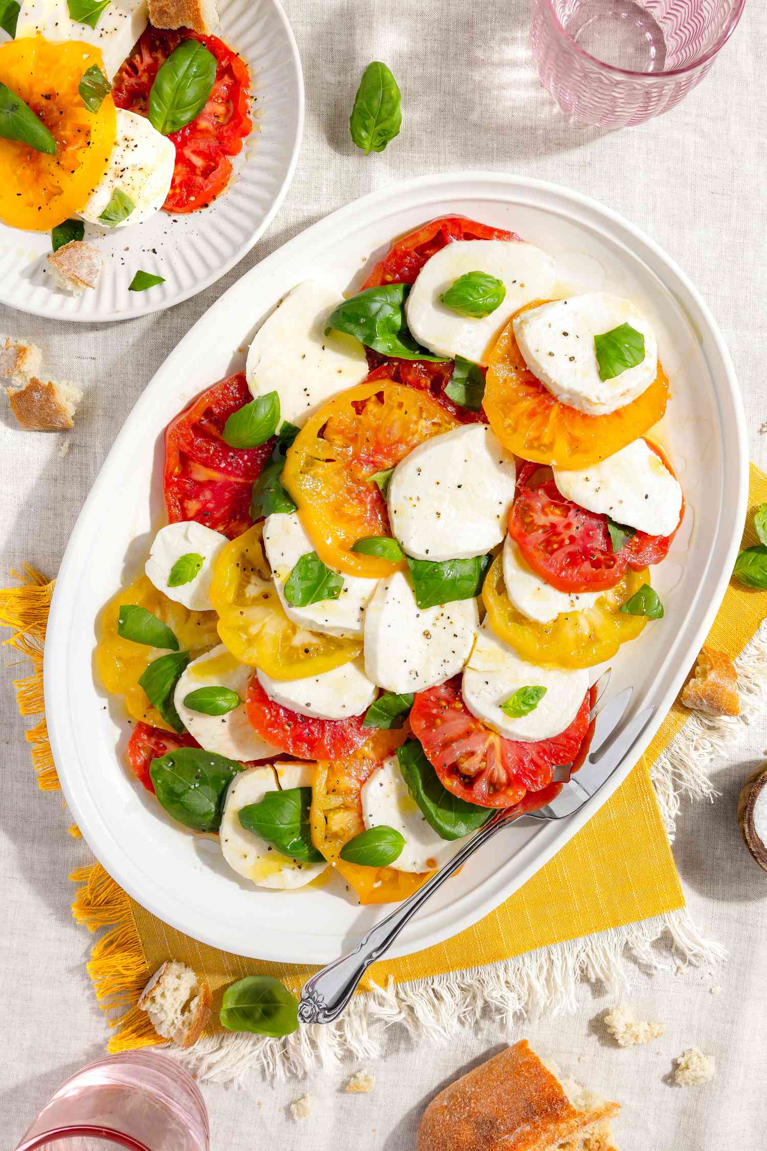 Table Setting With a Platter of Caprese Salad With Tomatoes, Basil, and Mozzarella All on a Yellow Placemat. a Plate With a Serving of Caprese Salad, Two Glasses of Water, Piece of a Baguette on the Table, and Small Bite Pieces on Various Parts of the Table