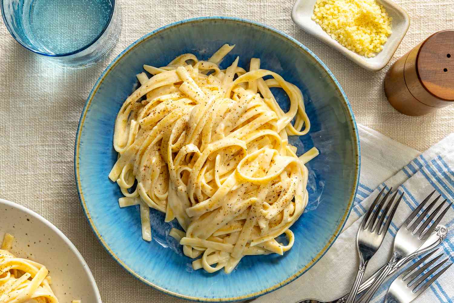 A plate of fettuccine Alfredo in a blue bowl, garnished with grated cheese on the side, with silverware and a napkin nearby