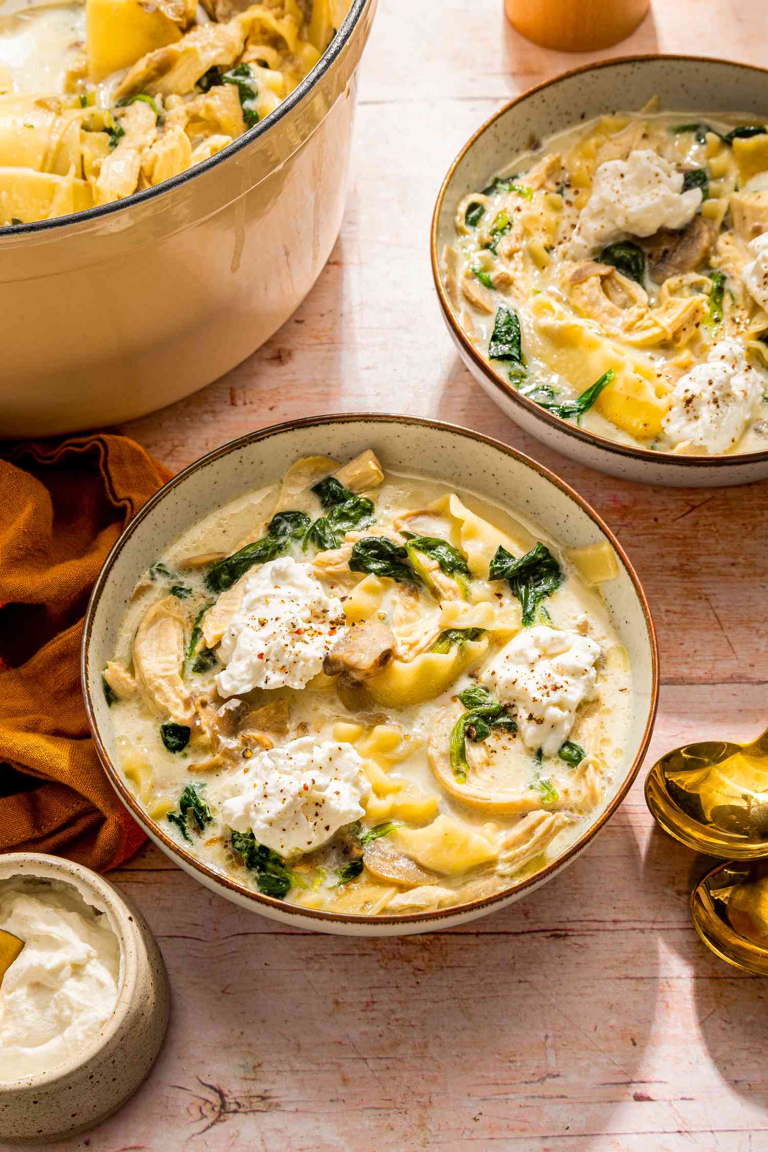 White chicken lasagna soup served in two bowls at a table setting with the spoons, napkins, and the rest of the soup in a dutch oven