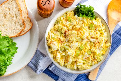 bowl of egg salad with a piece of lettuce on the counter next to a plate with bread and lettuce, salt and pepper shakers, and large spoon on a table napkin