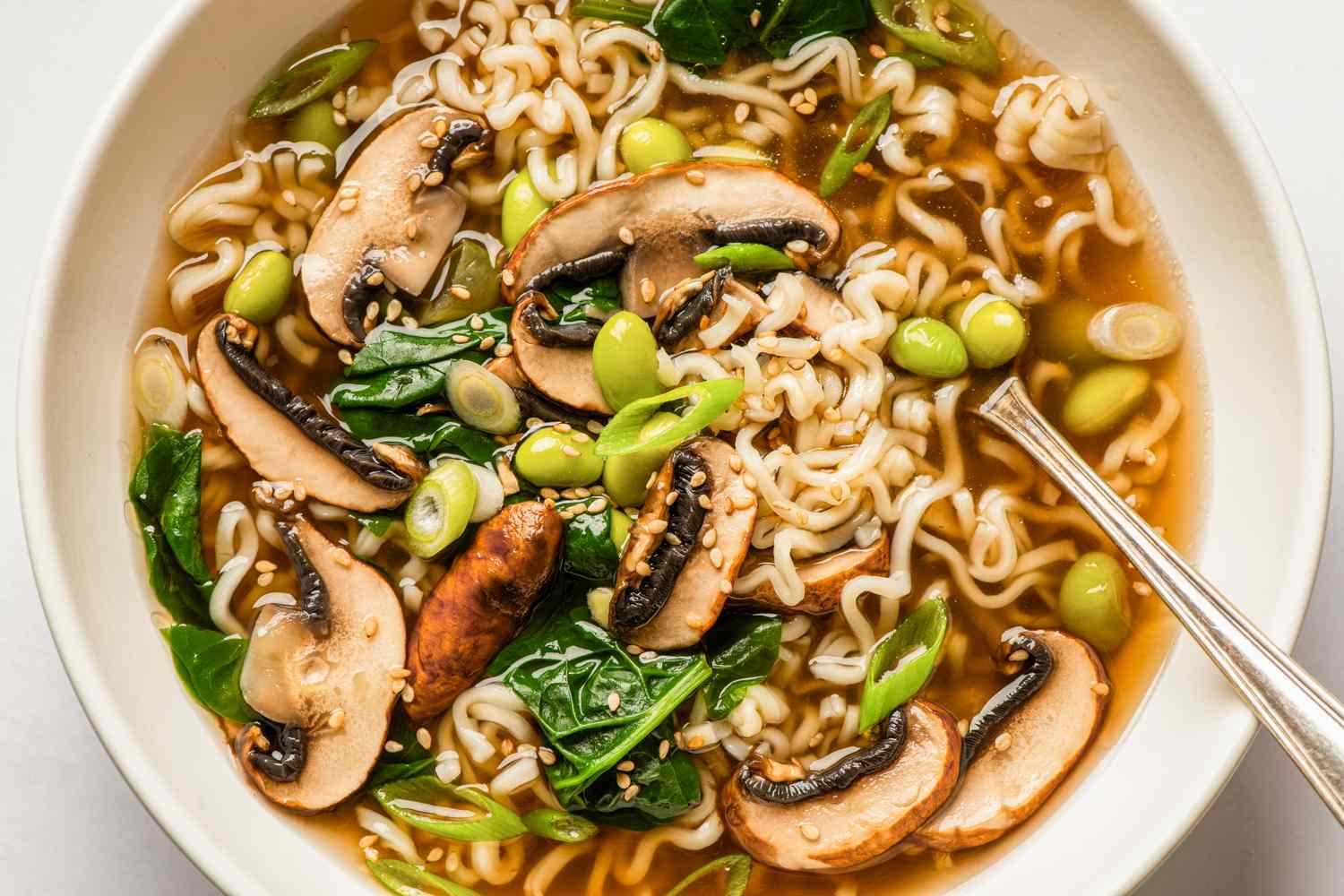 A bowl of noodle soup containing mushrooms spinach edamame green onions and sesame seeds with a spoon on the side