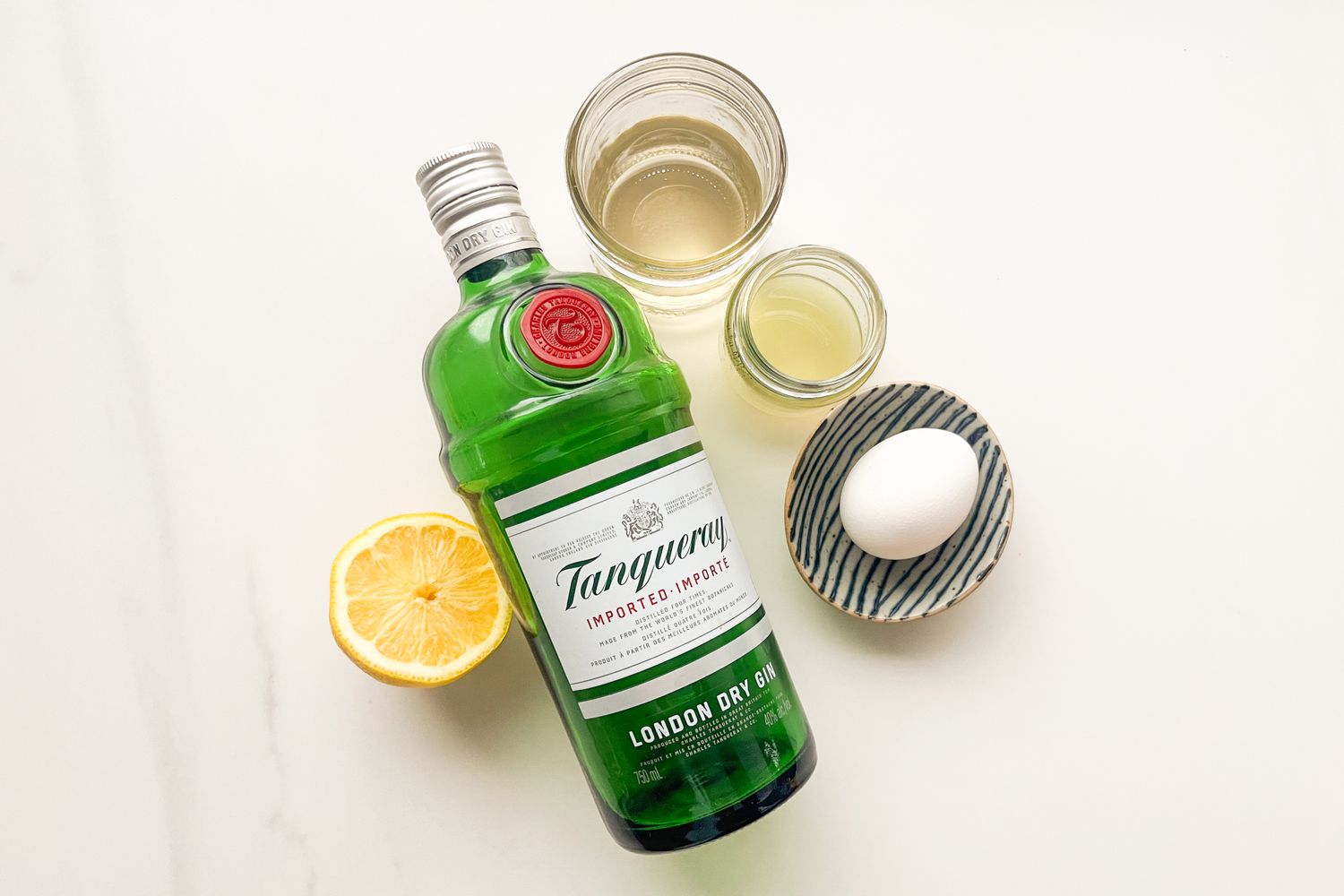 Ingredients for a cocktail on a table including a bottle of Tanqueray gin a lemon half an egg and two glasses with liquids
