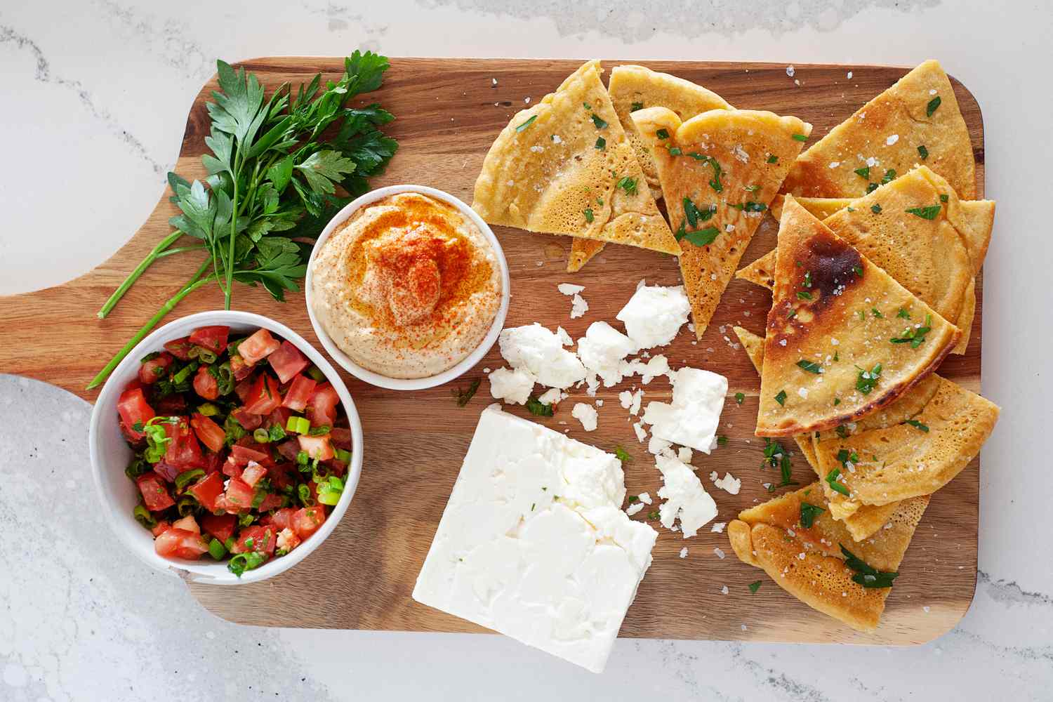 A wooden cutting board with slices of Chickpea Flour Flatbread, soft cheese, salsa, and hummus.