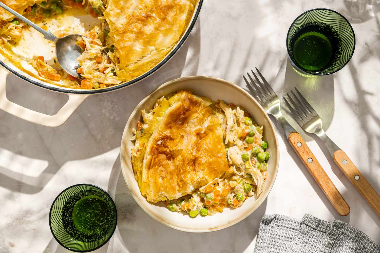 A serving of pot pie in a bowl next to a pot with a spoon on a table set with utensils and glasses