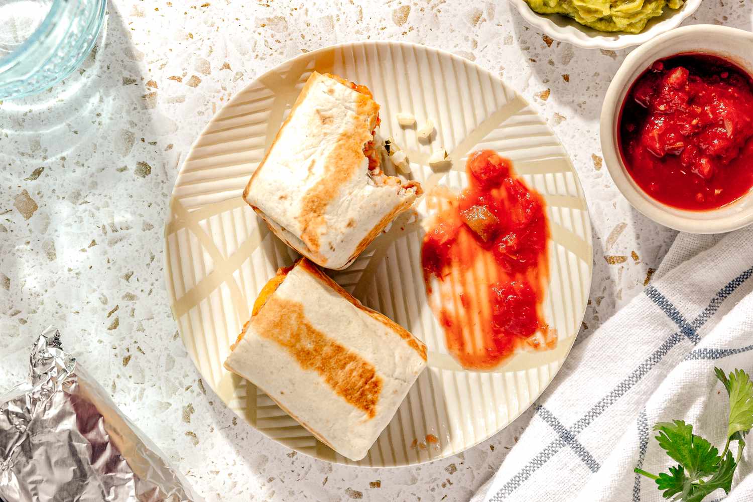 Easy Rice and Beans Freezer Burrito Cut in Half on a Plate and Served With Salsa, and in the Surroundings, a Small Bowl of Salsa, a Small Bowl of Guacamole, More Burritos Wrapped in Aluminum Foil, a Glass of Water, and a Kitchen Towel