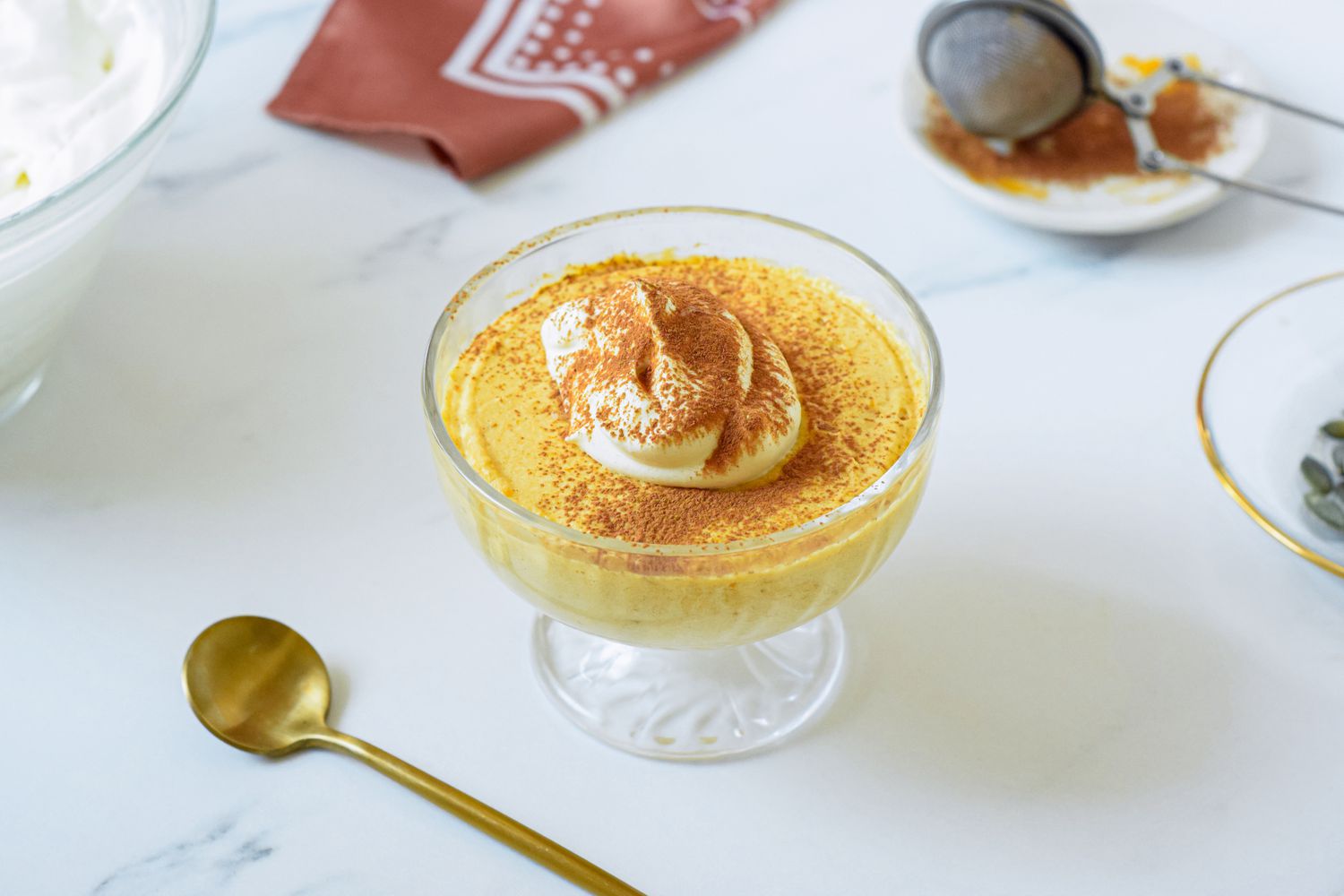 Single serving of 2-ingredient pumpkin mousse topped with whiped cream and a dusting of pumpkin spice in a sherbert glass, and in the surroundings, a bowl with more whipped cream, a bakers dusting wand filled with pumpkin spice on a small saucer, and a brown kitchen linen